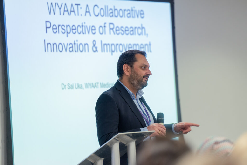 Dr Sal Uka presenting on WYAAT stood in front of a presentation slide on "WYAAT: A Collaborative Perspective of Research, Innovation & Improvement