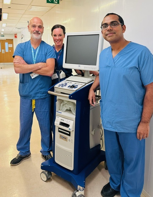 An image of the team with 3 of the team wearing medical scrubs and uniforms stood with a piece of medical monitoring equipment.