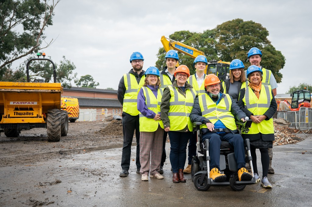 Group of nine people in hard hats and high-vis jackets standing in front of a dump truck on a building site. One person is seated in a wheelchair