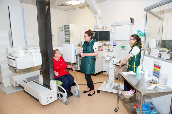 The photograph shows a patient sitting in the chair to have the video swallow, and also a speech and language therapist and a radiographer.