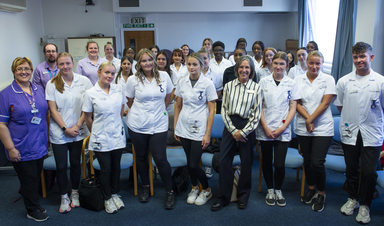 Large group of students dressed in clinical uniform and woman dressed in business clothing stand in a classroom