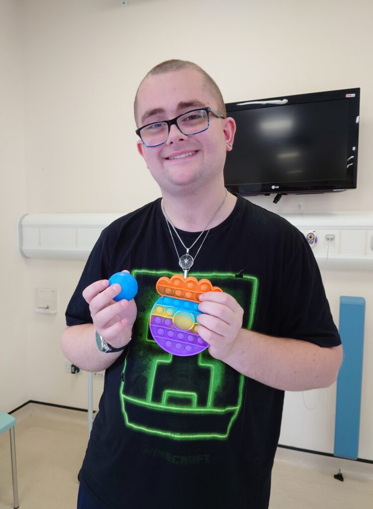 A young man in a black t-shirt and glasses holding a blue stress ball and fidget toy.