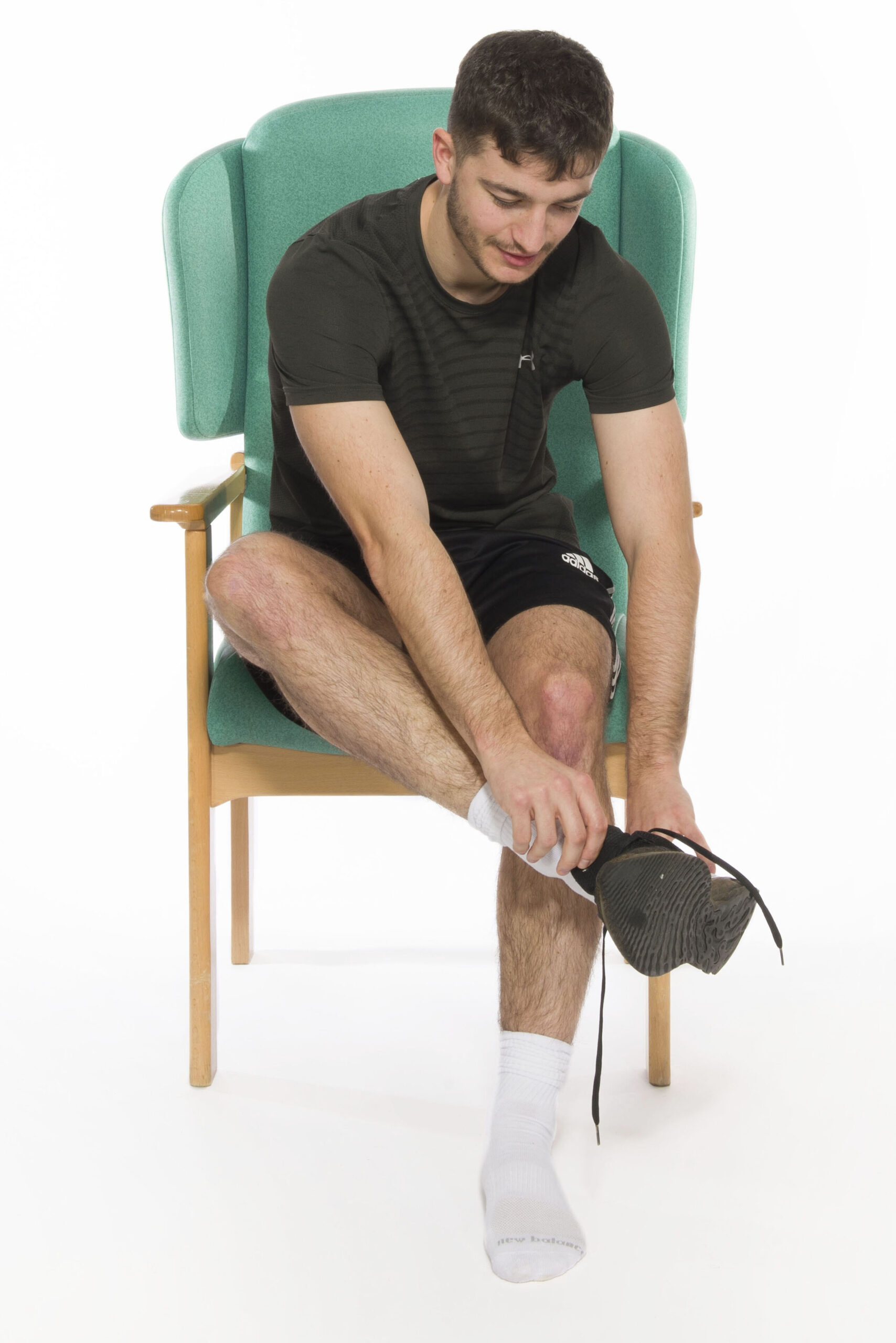 Photograph of a person sat in a chair lifting their foot up to put a shoe on.