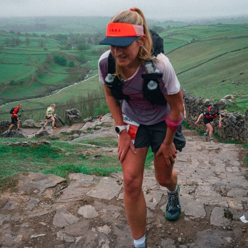 Mel Sykes, wearing shorts and running vest and visor, scaling a steep hill with other runners behind her. 