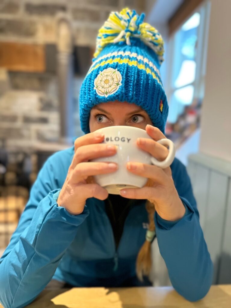 Mel, wearing a blue and yellow woolly hat and a blue running jacket, holding a cup of tea. 