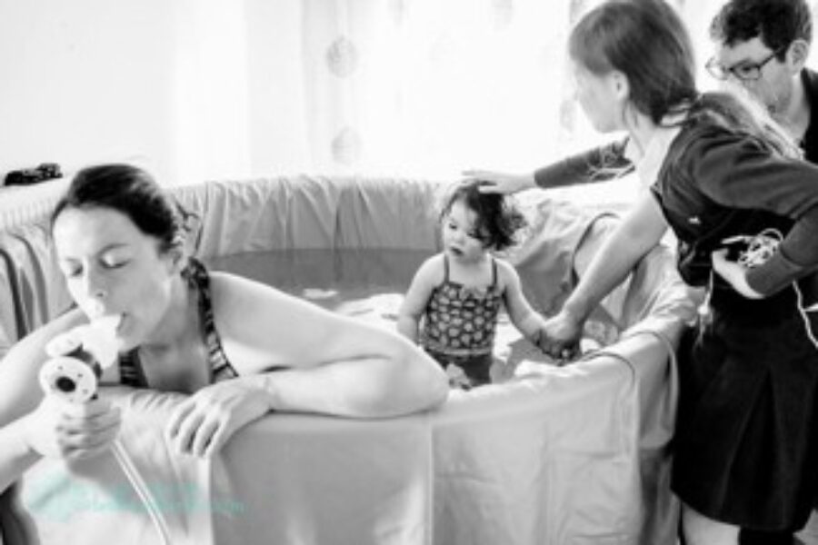 Black and white photo of mum in labour in birthing pool using gas and air. Toddler is stood in pool and dad and older daughter stood at the side.