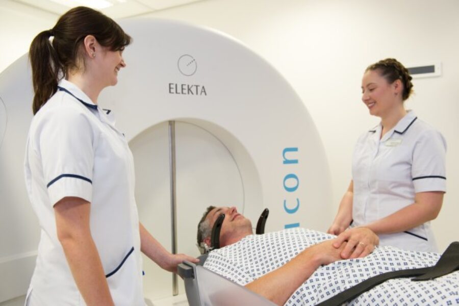 Two people in clinical uniforms standing beside a person lying down, next to a white spherical diagnostic machine