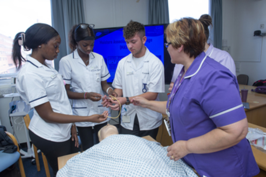 People dressed in clinical uniforms practice with a piece of medical equipment on a dummy patient which is lying on a table