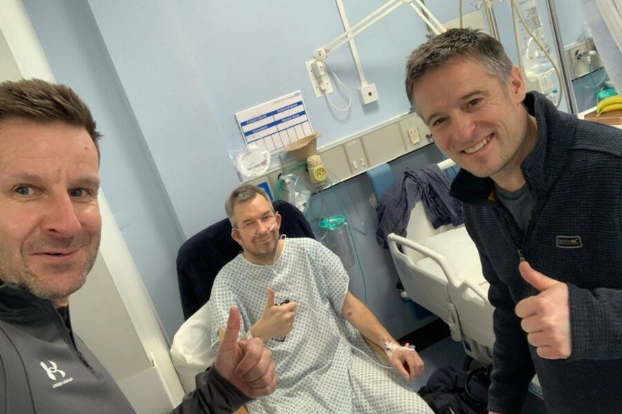 Mark in a hospital gown in a chair. Mark, Gareth and another friend putting up their thumbs smiling.