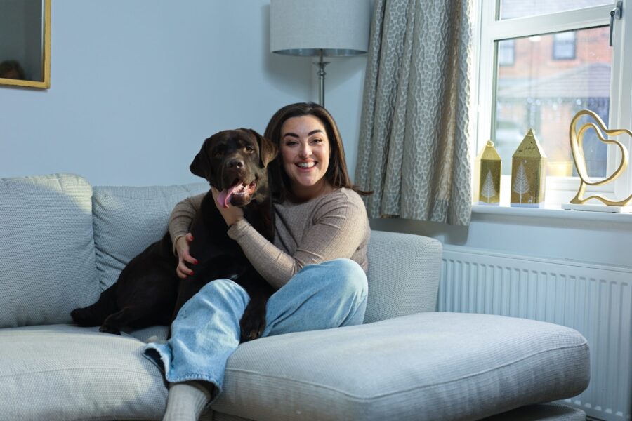 A young woman sat on a grey sofa in a living room cuddling a brown dog.